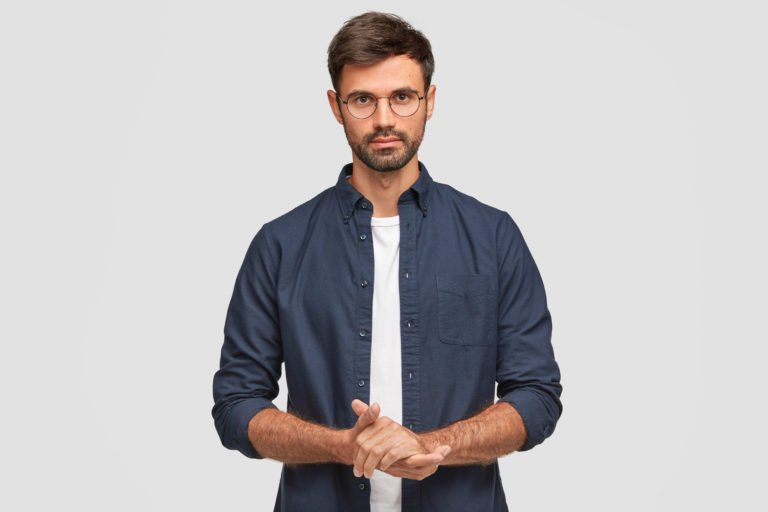 Waist up portrait of handsome serious unshaven male keeps hands together, dressed in dark blue shirt, has talk with interlocutor, stands against white background